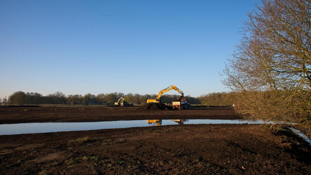 Graafmachines werken op bouwterrein bij waterplas in open landschap onder blauwe lucht
